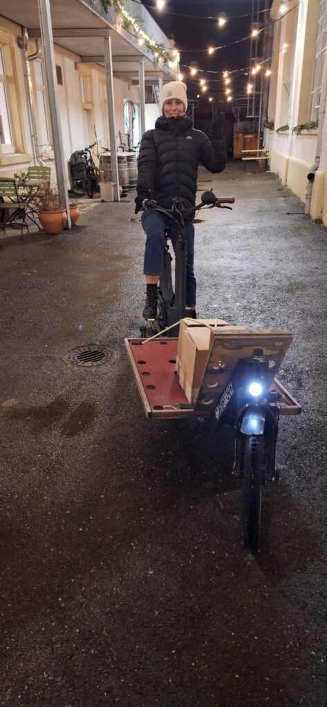 Bild 1: Lastenrad-Lieferung ALT: Frau auf einem Lastenrad mit Paket in einem abendlichen Innenhof unter Lichterketten.