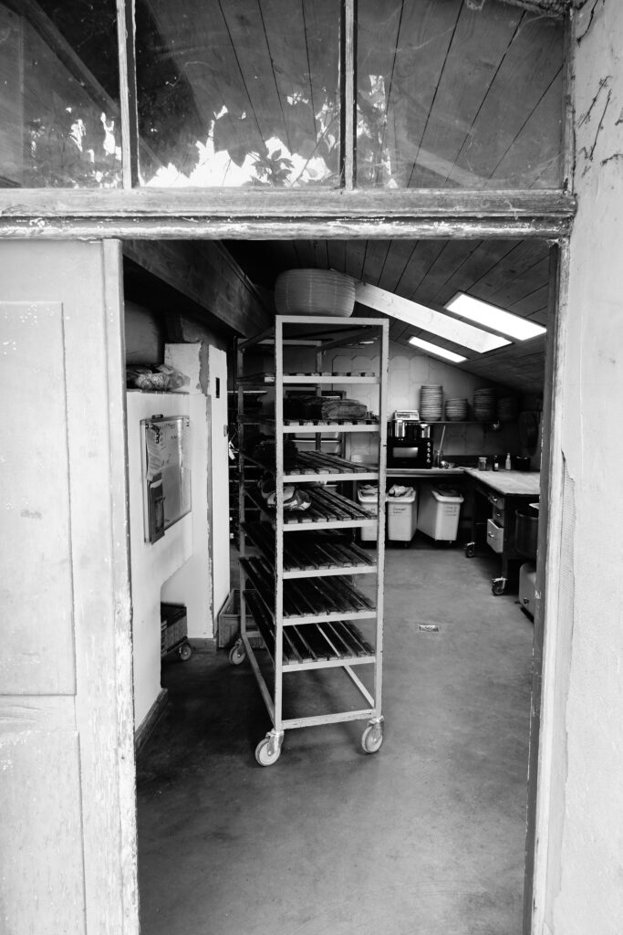 Blick hinter die Kulissen: Eine geöffnete Tür zeigt die helle, funktionale Backstube des Backhauses der Vielfalt mit Arbeitsflächen, Teigwannen und einem Regalwagen.