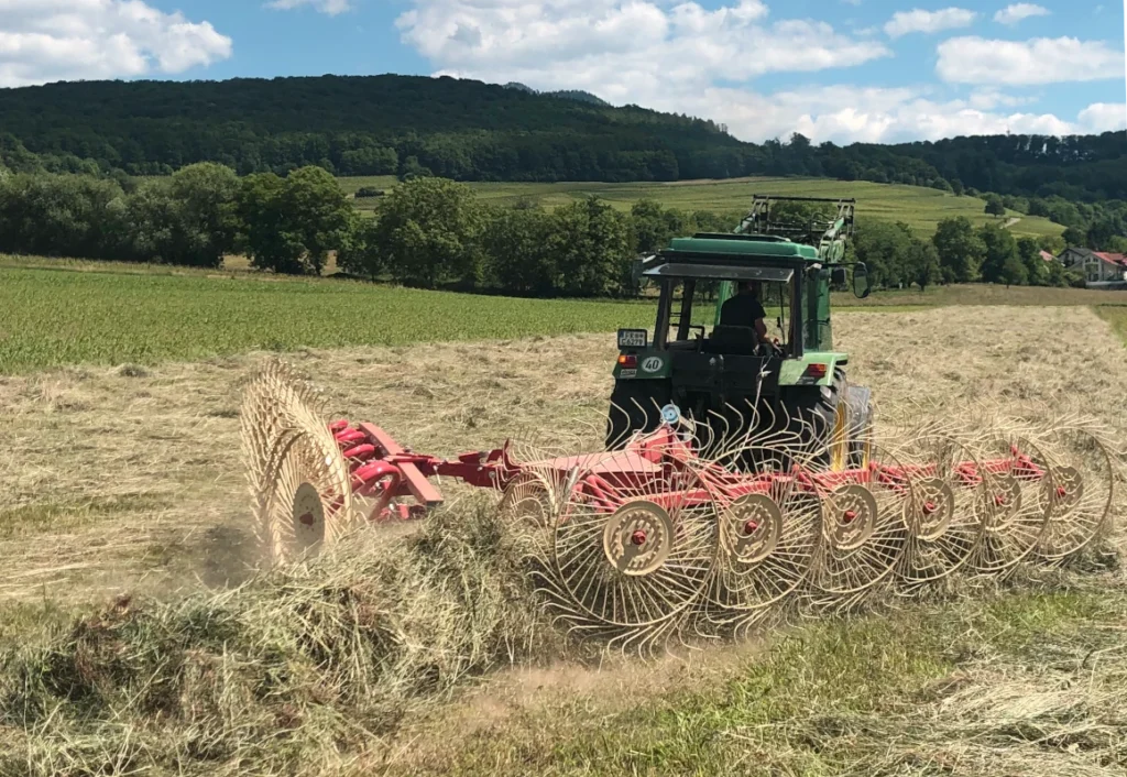 Futter für den Hofkreislauf: Ein Traktor beim Heu-Schwaden auf den weiten Wiesen der Solawi Luzernenhof vor der grünen Hügelkulisse.