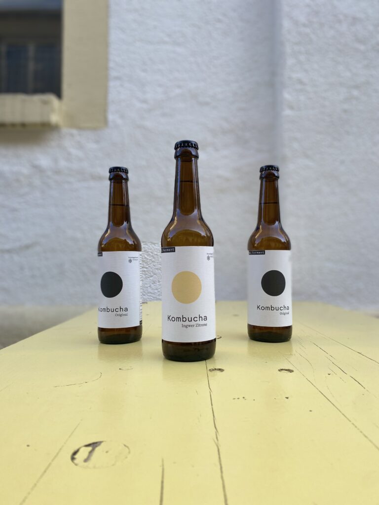 Drei Flaschen St. Ferment Kombucha stehen auf einem hellgelben Holztisch, in der Mitte die Sorte Ingwer Zitrone, flankiert von zwei Flaschen der Sorte Original.
