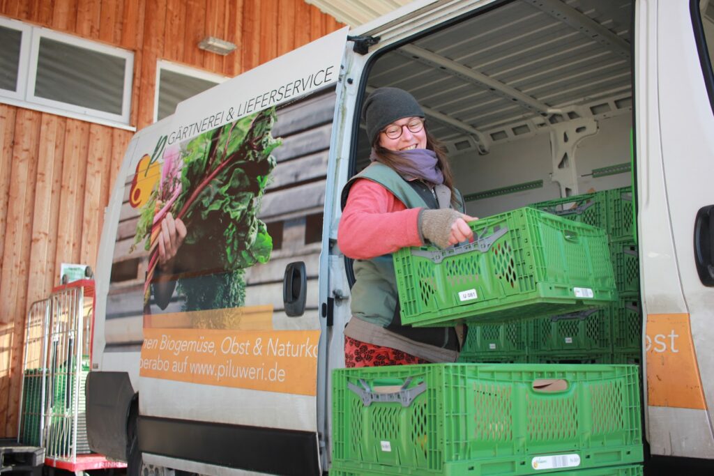Fröhliche Piluweri-Mitarbeiterin belädt den Lieferwagen mit einer grünen Kiste für die Bio-Abokiste und den Lieferservice.
