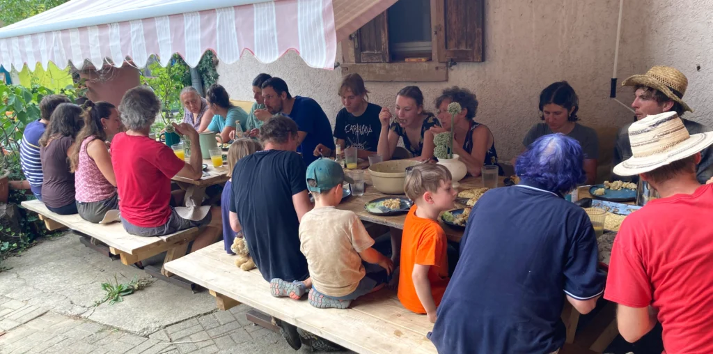 Gelebte Gemeinschaft: Viele Erntehelfer und Mitglieder sitzen an einer langen Tafel im Freien beim gemeinsamen Mittagessen auf dem Luzernenhof zusammen.