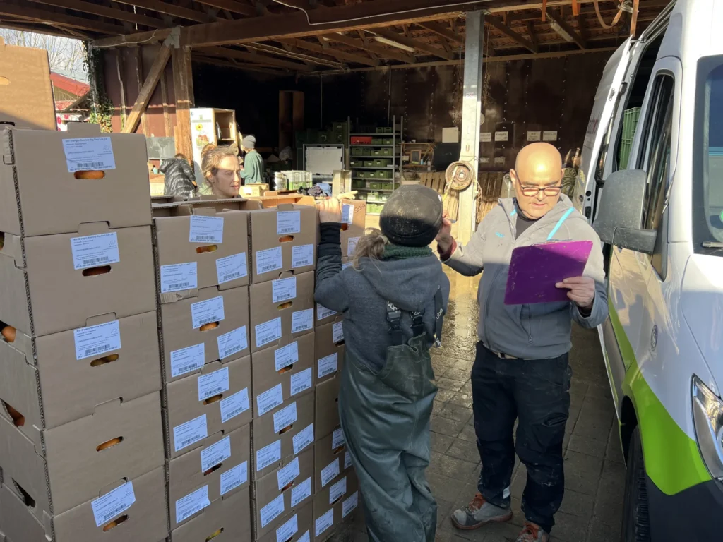 Logistik auf dem Hof: Koordination der Anlieferung – ein Koordinator mit Klemmbrett überprüft die Paletten mit Orangenkisten, während das Team beim Entladen des Stadtmobil-Transporters hilft.