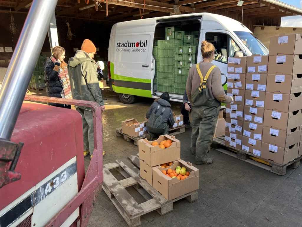 Solidarische Logistik: Ein Stadtmobil-Transporter wird auf dem Hof entladen – Mitglieder und Landwirte organisieren gemeinsam die Ankunft der großen Stapel mit griechischen Orangenkisten.