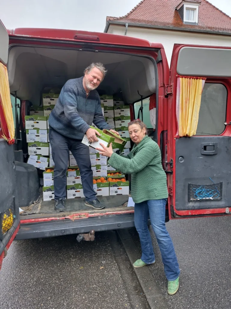 Einsatz für die Gemeinschaft: Zwei engagierte Mitglieder entladen bei Regenwetter gut gelaunt grüne Teikei-Kisten aus einem roten Lieferwagen für die lokale Verteilung.