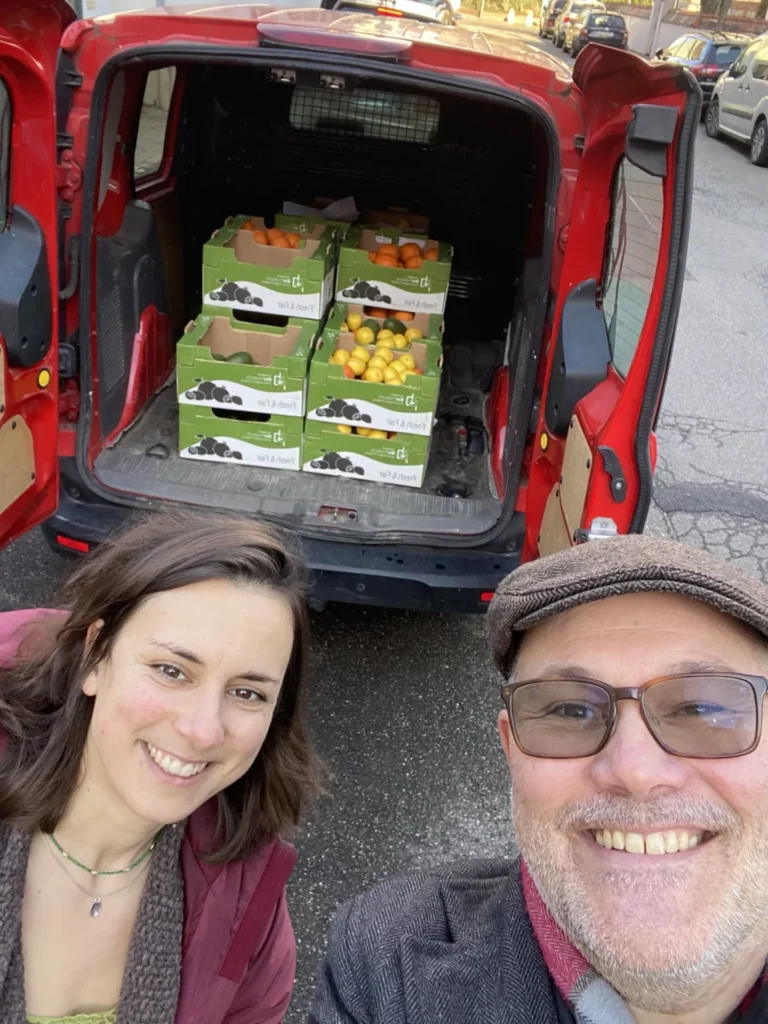 Glückliche Gesichter bei der Ankunft: Zwei Mitglieder der Teikei-Community stehen strahlend vor einem Lieferwagen voller frischer Orangen-Kisten bei einer Verteilaktion in Deutschland.