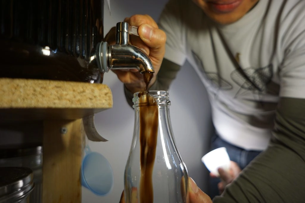 Zero Waste im Alltag: Ein lächelndes Mitglied der FoodCoop L13 zapft frische flüssige Lebensmittel aus einem großen Glasspender direkt in eine mitgebrachte Mehrwegflasche.