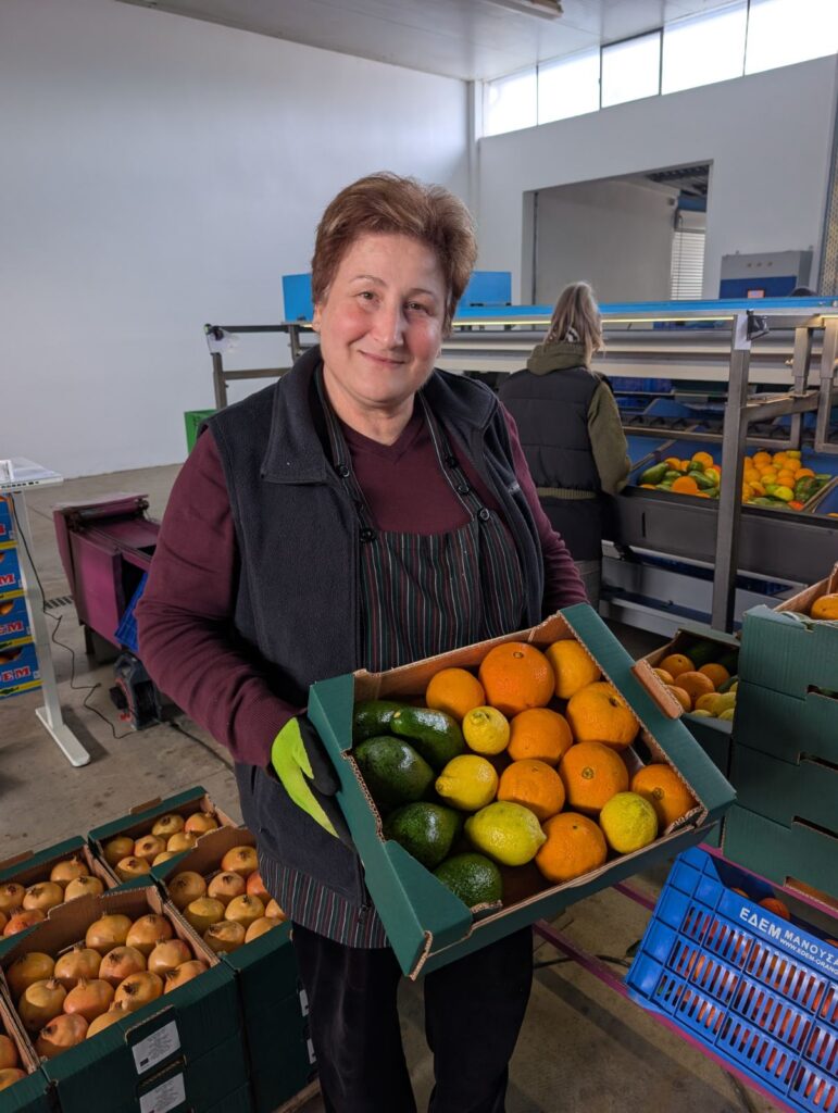 Persönlicher Bezug zum Produkt: Eine stolze Landwirtin auf Kreta präsentiert lächelnd eine frisch gepackte Teikei-Mischkiste mit Bio-Orangen, Zitronen und Avocados.