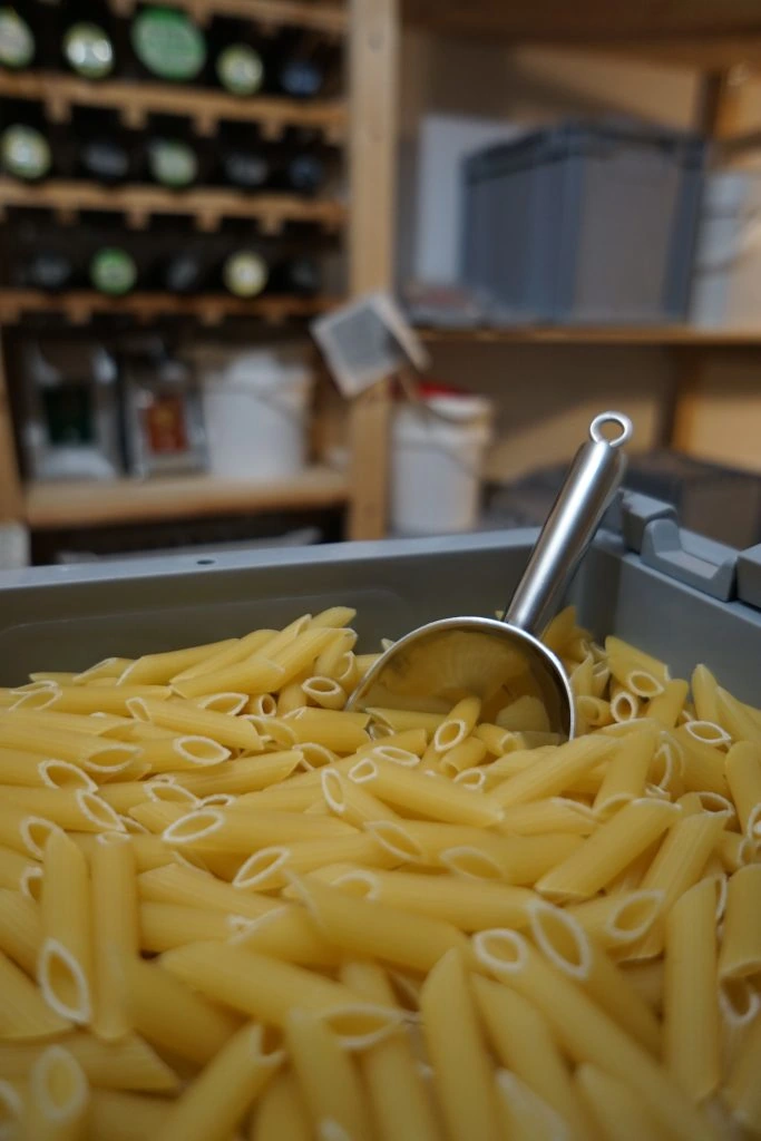 Verpackungsfreie Lebensmittel: Nahaufnahme einer Vorratskiste voller Penne-Nudeln mit einer Metallschaufel, bereit zur individuellen Abfüllung im Regal der FoodCoop L13.