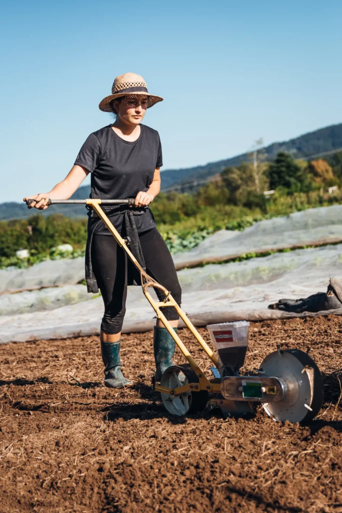 Frühlingsarbeit auf dem Acker: Eine Gärtnerin mit Strohhut schiebt eine manuelle Sämaschine über das Feld des Lebensgarten Dreisamtal, um neues Gemüse auszusäen.