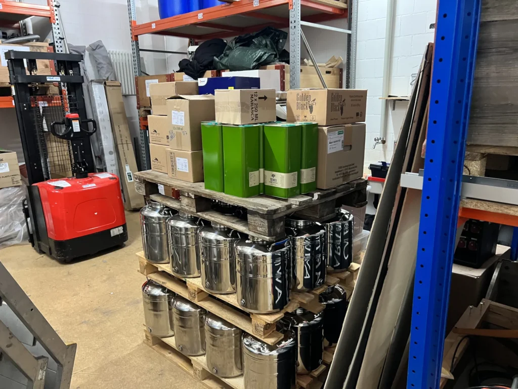 Logistik für faires Olivenöl: Eine Palette mit großen Edelstahl-Behältern und grünen 5-Liter-Kanistern voller Teikei-Olivenöl steht im Lager bereit für die Verteilung.