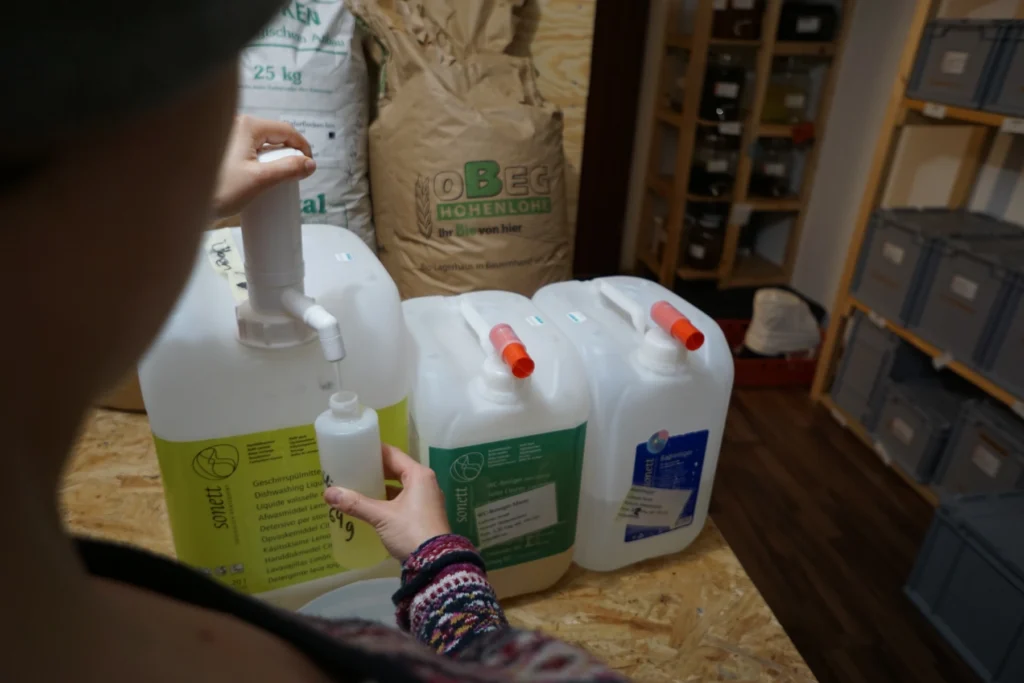 Nachhaltig einkaufen: Eine Frau füllt in der FoodCoop L13 ökologisches Spülmittel aus einem großen Nachfüllkanister direkt in eine eigene, wiederverwendbare Flasche ab.