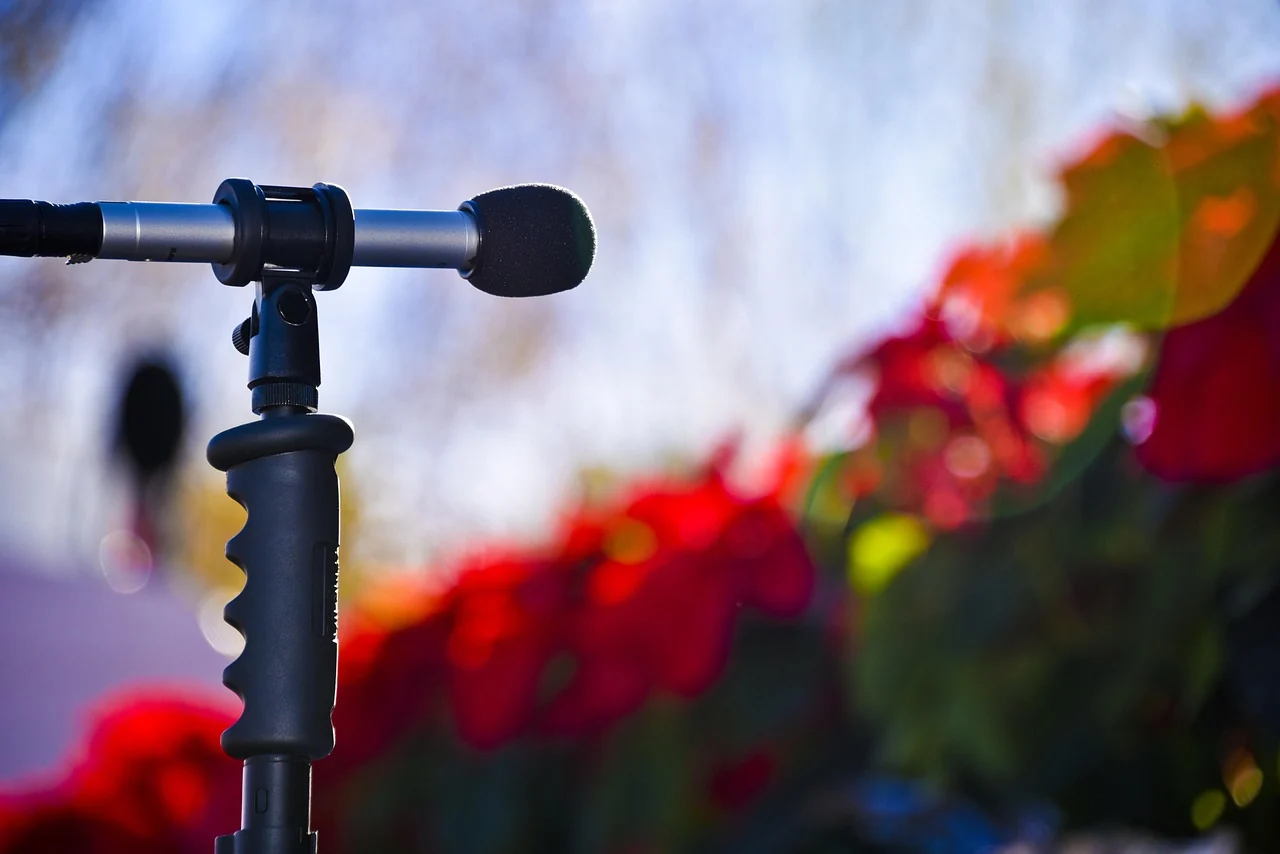 Professionelle Kommunikation und Moderation – KEWI Freiburg Nahaufnahme eines Mikrofons auf einem Stativ vor einem unscharfen Hintergrund mit roten Blumen im Freien.